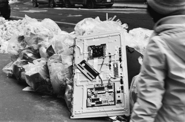 a white metal sheet with attached boards and wiring rests, discarded, against white trash bags and snow piled up on a new york city sidewalk