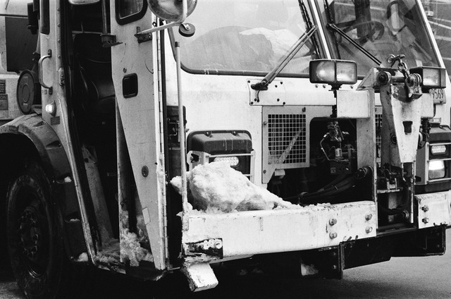 close up view of the front and a bit of the side of a nyc garbage truck. the truck is partially covered in snow and looks particularly brutalist in the black and white rendering