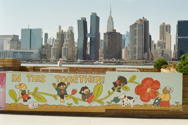 a picture of a cartoon mural of a diverse cast of kids playing on a big flower tilted sideways with an older lady with a walker smelling the flower contentedly. a cat basks under a leaf as a dog uses a watering can to nurture a seedling. behind the mural, in the background, is the new york skyline as seen from long island city.