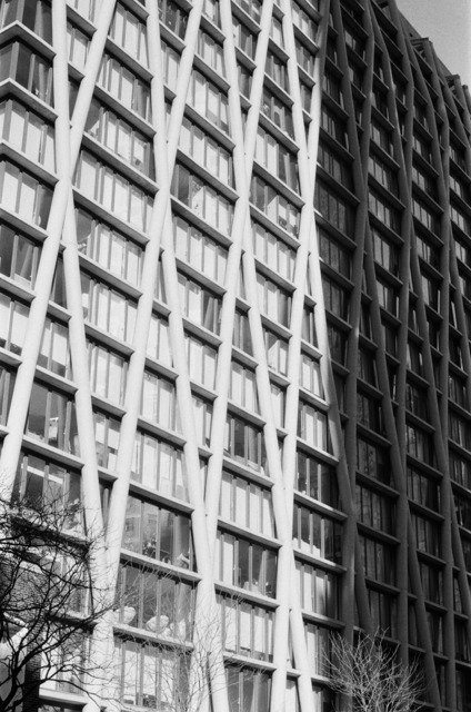 a building with prominent exterior triangular supports lies partly in shadow and partly in direct sunlight. the contrast is captured starkly in this black and white over-exposed photo